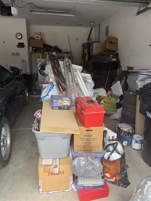 Cluttered garage with stacked boxes, tools, car parts, and miscellaneous items on work surfaces