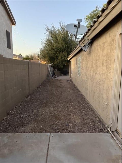Narrow residential side yard with gravel ground, tan block walls, fence, and satellite dish mounted on roof