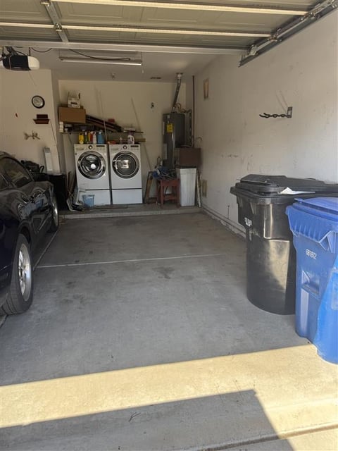 Garage interior with front portion of car visible on left, white washer and dryer units in center back, trash cans on right side, and storage items along walls