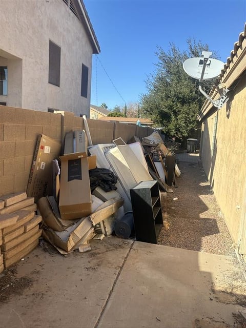 Cluttered backyard alley with stacked cardboard boxes, discarded furniture, and appliances near concrete block walls under clear blue sky